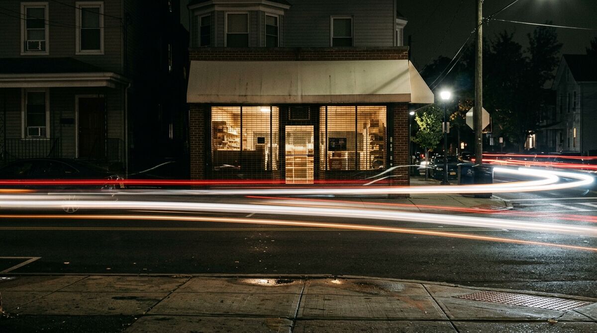 Corner shop at night with lit interior and long-exposure light trails of passing traffic streaking across the street in front